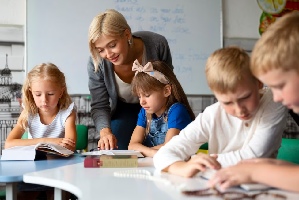 woman leans towards children in classroom and checks the notebooks